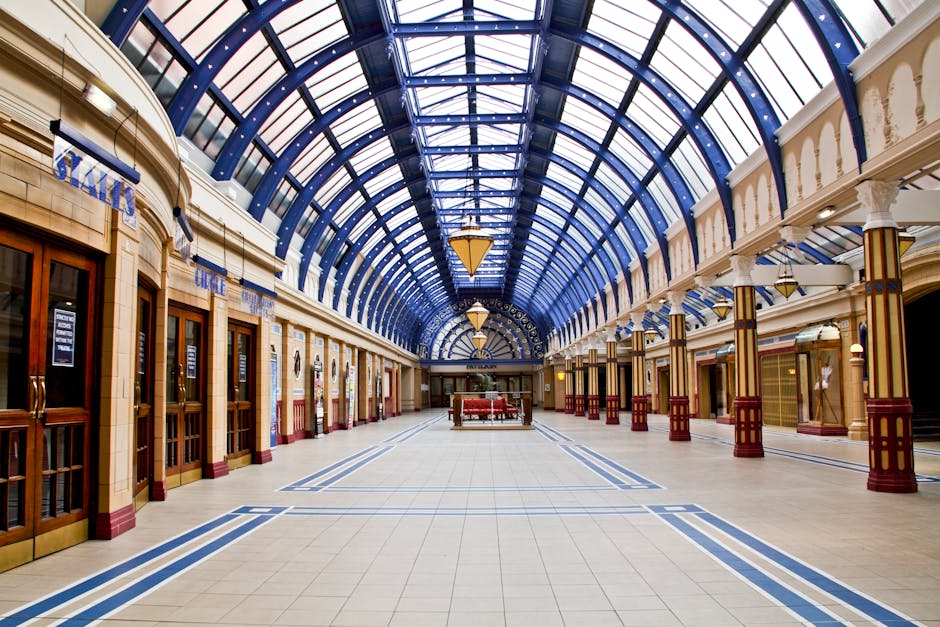 A spacious, well-lit indoor shopping arcade with a high, arched glass ceiling supported by ornate columns. The smooth tiled floor is clean and free of debris, featuring blue line patterns running alon